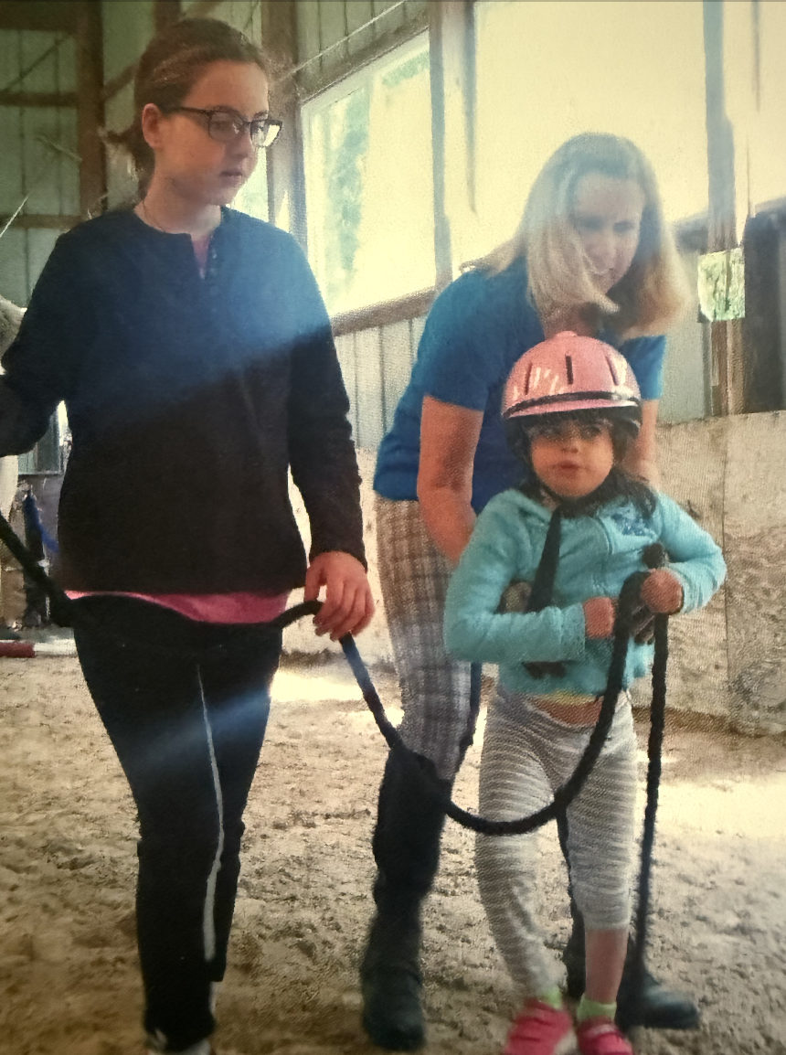 A young girl wearing a pink helmet and gloves, holding a harness, being led by two women inside a barn or riding arena with sunlight coming through the windows.