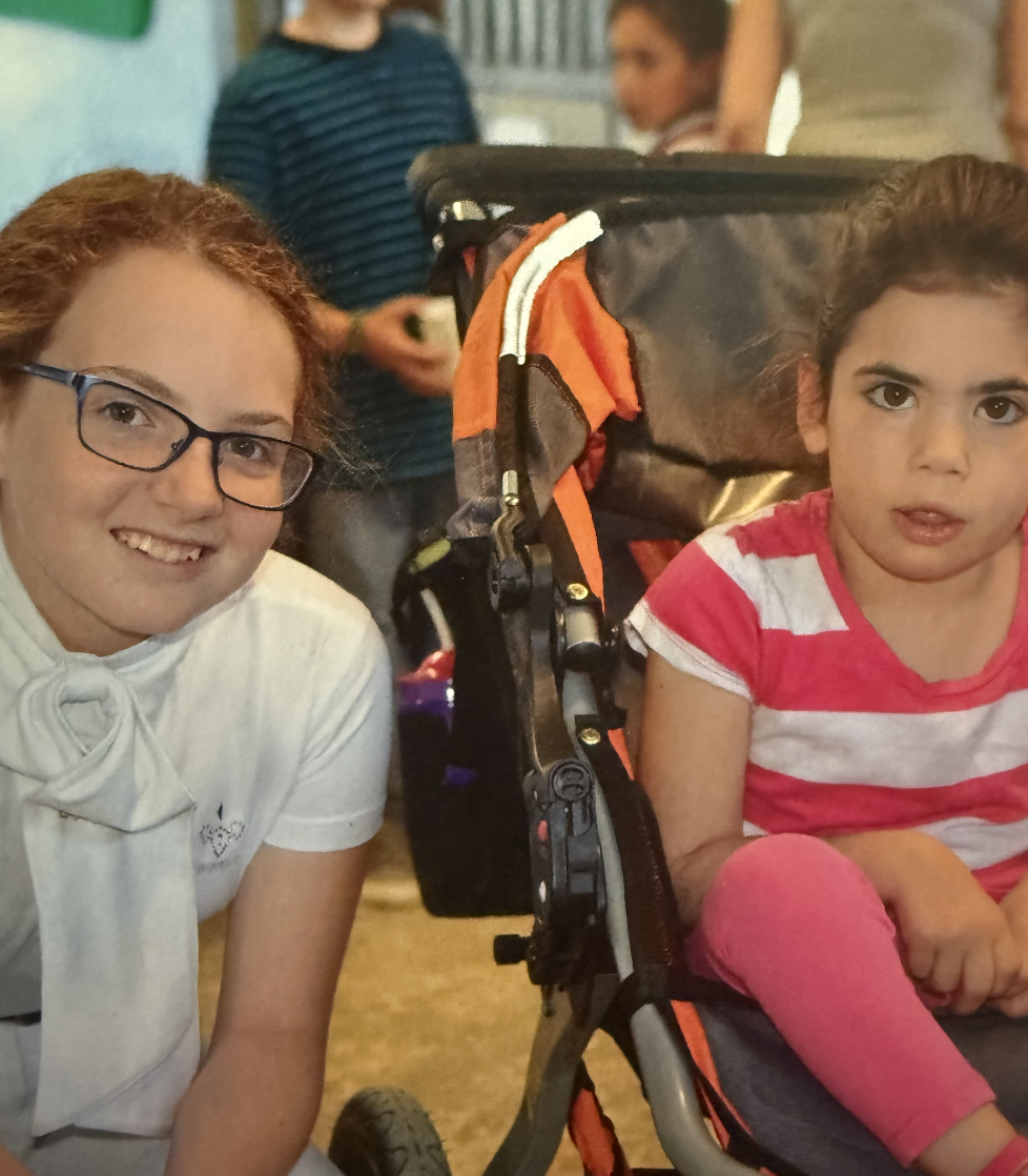 Two young girls sitting indoors, one smiling and wearing glasses, the other with a serious expression, sitting in a stroller. A woman stands in the background.