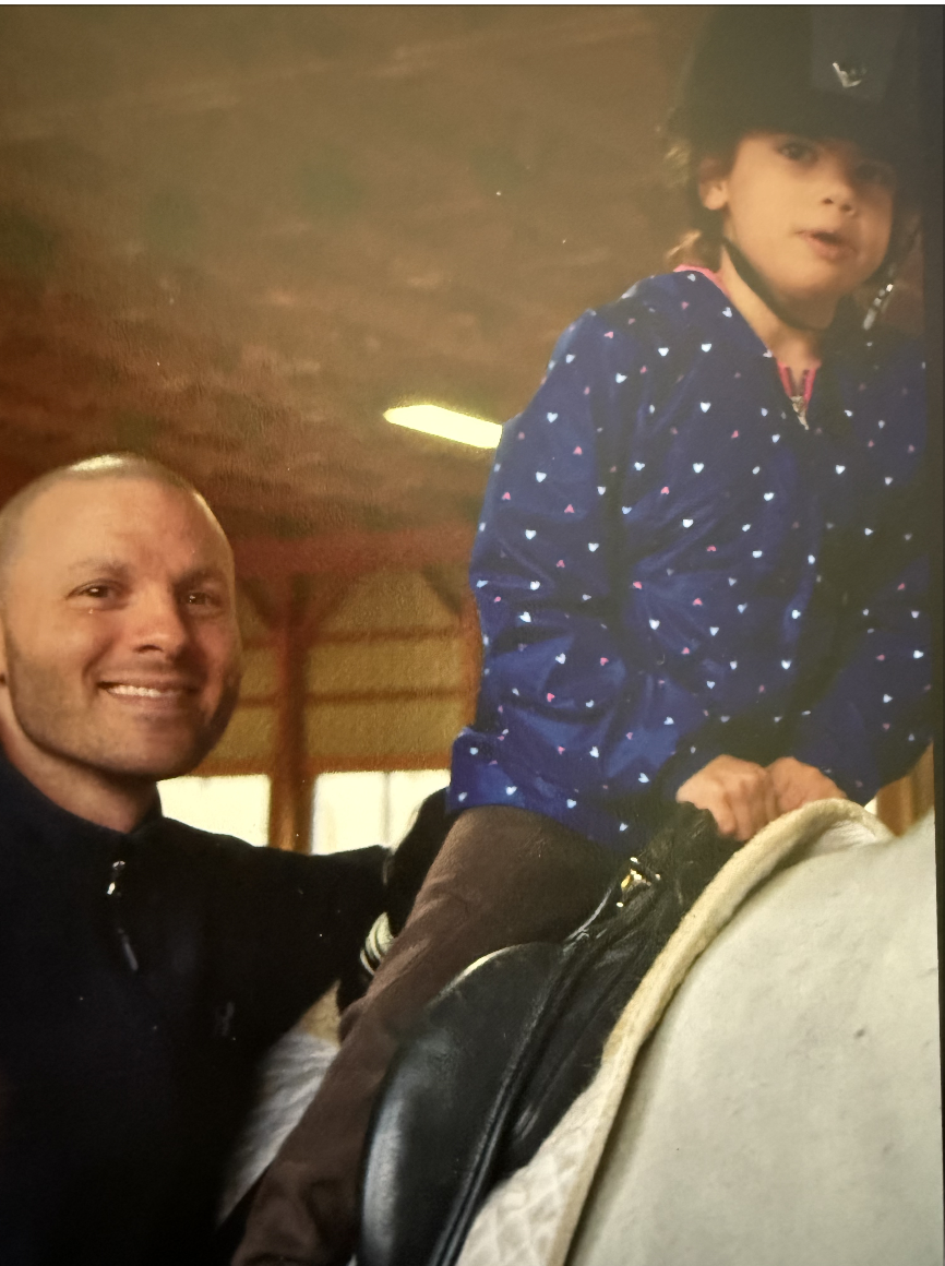 A man and a young girl riding a carousel horse, with the man smiling directly at the camera. The girl is sitting on the horse, holding the reins, and wearing a blue jacket with a star pattern, while the man is standing next to her.