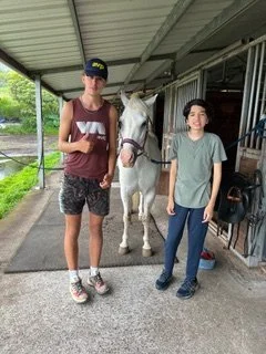 Two children and a white horse standing under a shelter at a stable or farm, with greenery in the background.