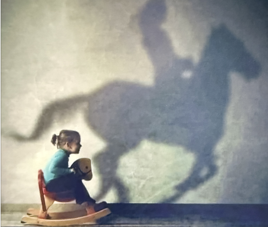 Child sitting on a rocking horse, casting a shadow of a larger horse on the wall.