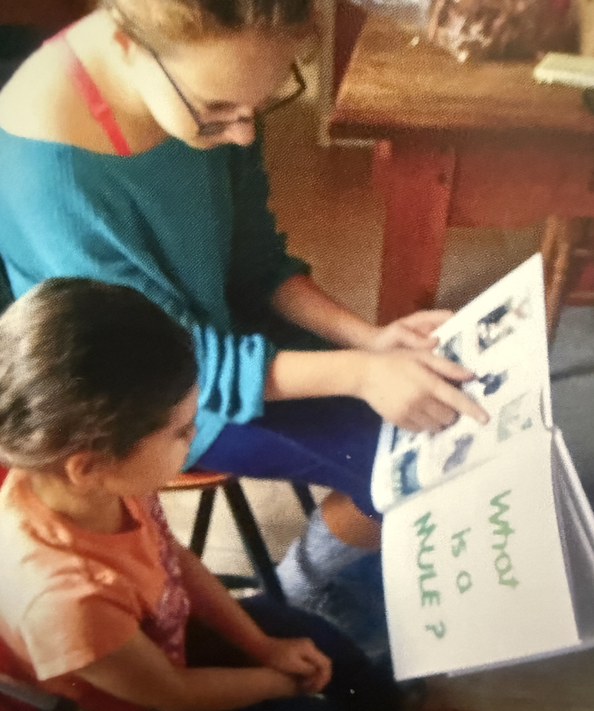 Two children sitting on chairs, looking at and pointing to a book or photo album with handwritten text that says 'WHERE'S DAD AND MOM'.