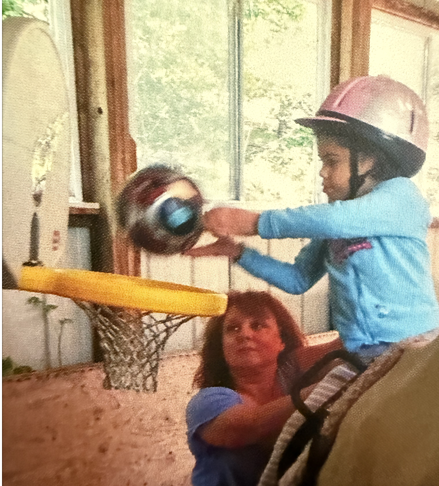 A young girl wearing a pink helmet and blue long sleeve shirt is hitting a basketball into a small indoor basketball hoop, while a woman with curly hair supervises nearby.