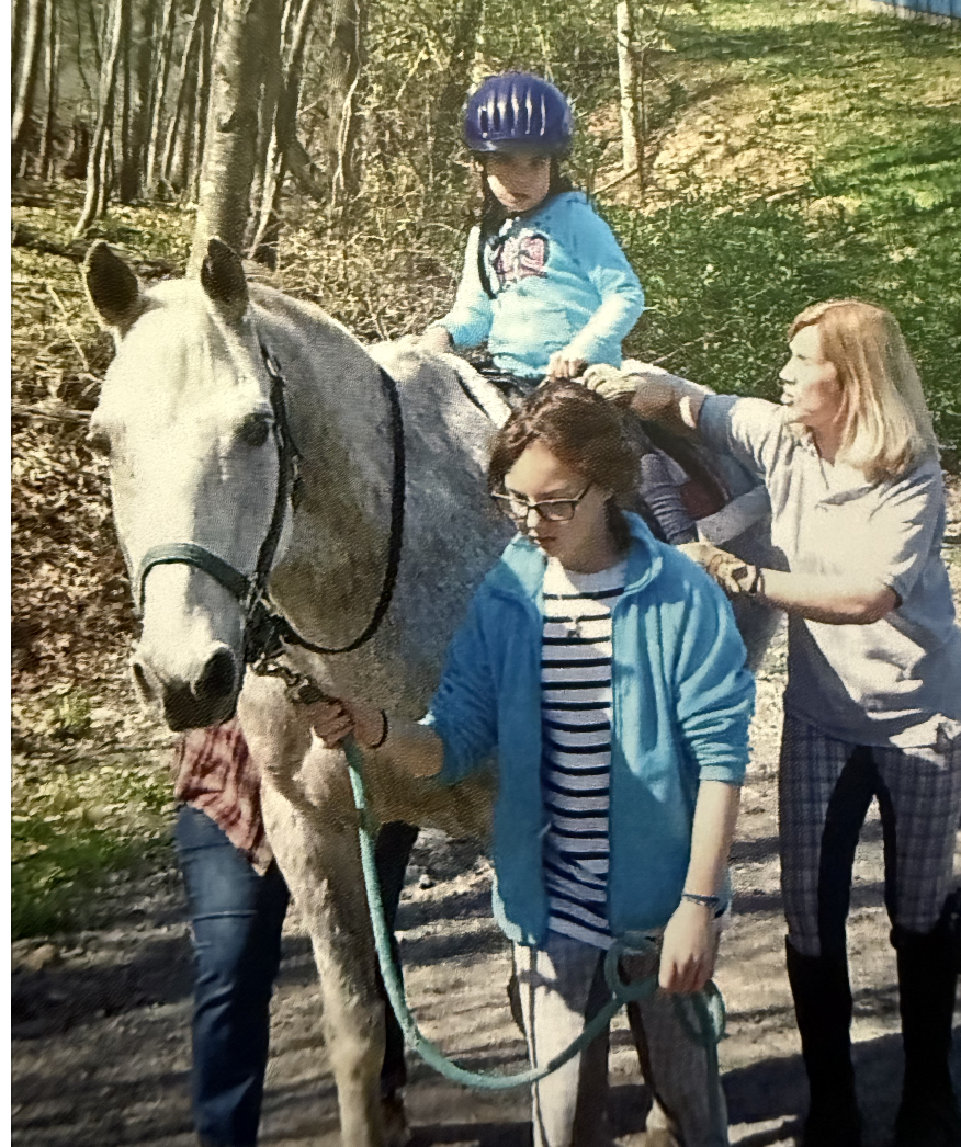 A young girl wearing a blue helmet and a light blue jacket rides a white horse with help from two women, one holding the horse's reins and the other assisting the girl on the horse, in a wooded area.