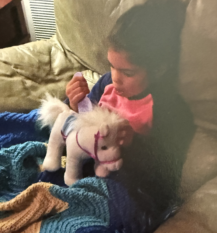 A young girl with dark hair sitting on a sofa, holding a plush toy horse and looking at it. The girl is wearing a pink shirt and a blue jacket, and there are blankets on her lap.