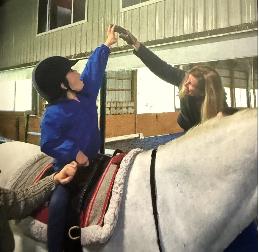 A young boy in a blue jacket and helmet giving a high five to a woman on horseback in an indoor riding arena.