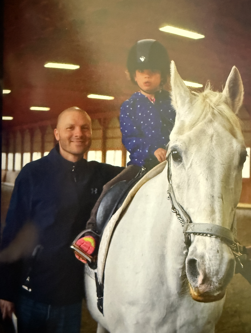 A young girl sitting on a white horse with a man standing next to her inside an indoor riding arena.