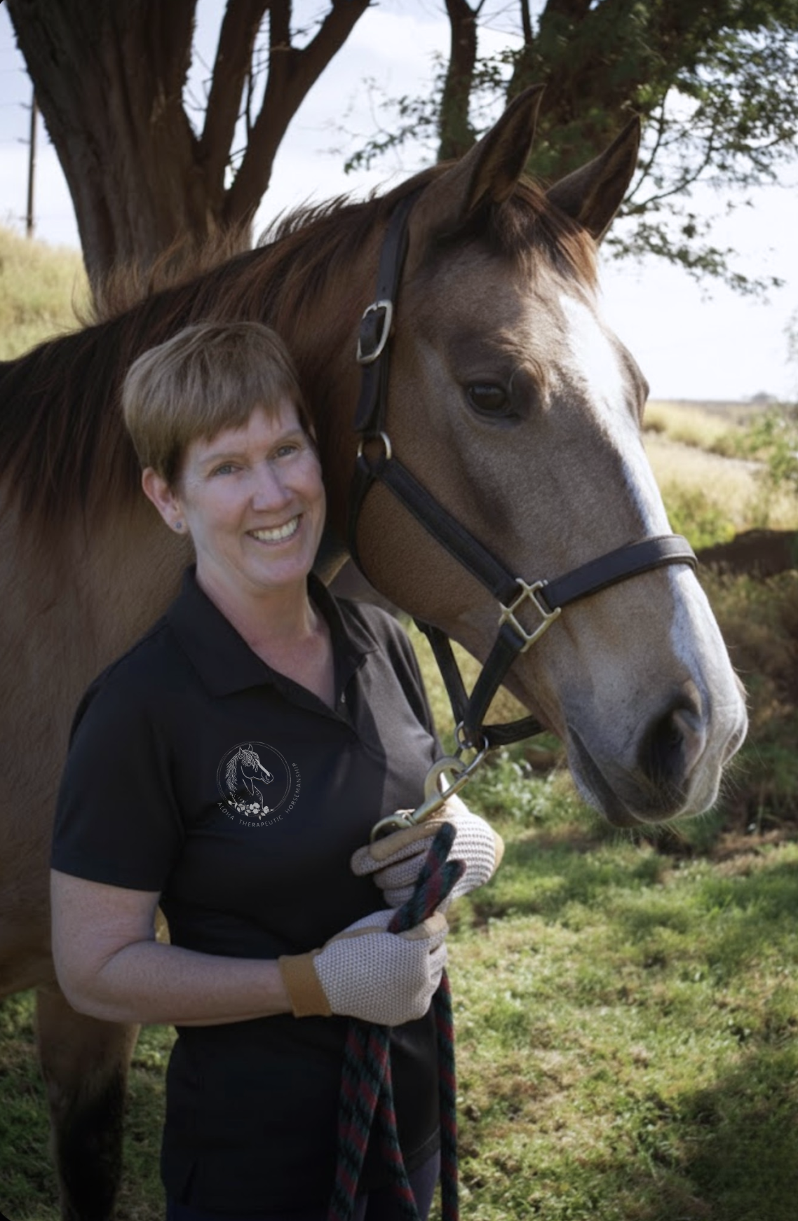 A smiling woman with short hair standing outdoors next to a brown horse, holding a lead rope.