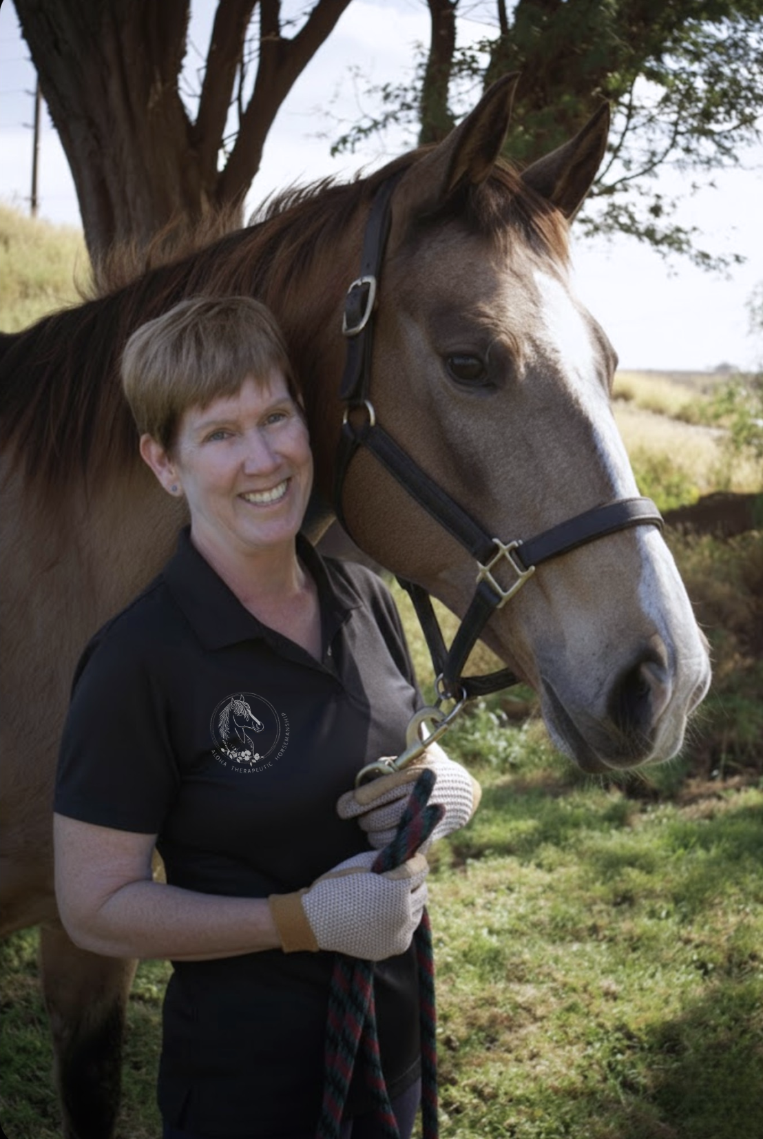 A woman standing in a grassy field holding a horse's lead rope, smiling at the camera with a large tree and open landscape in the background.