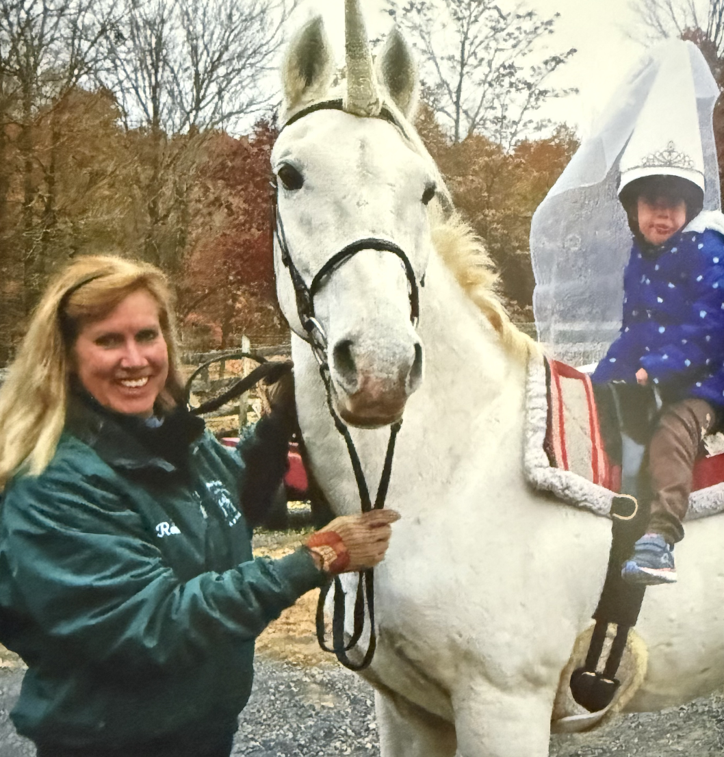 A woman smiling and holding a white horse with a child riding on it, outdoors with trees in the background.