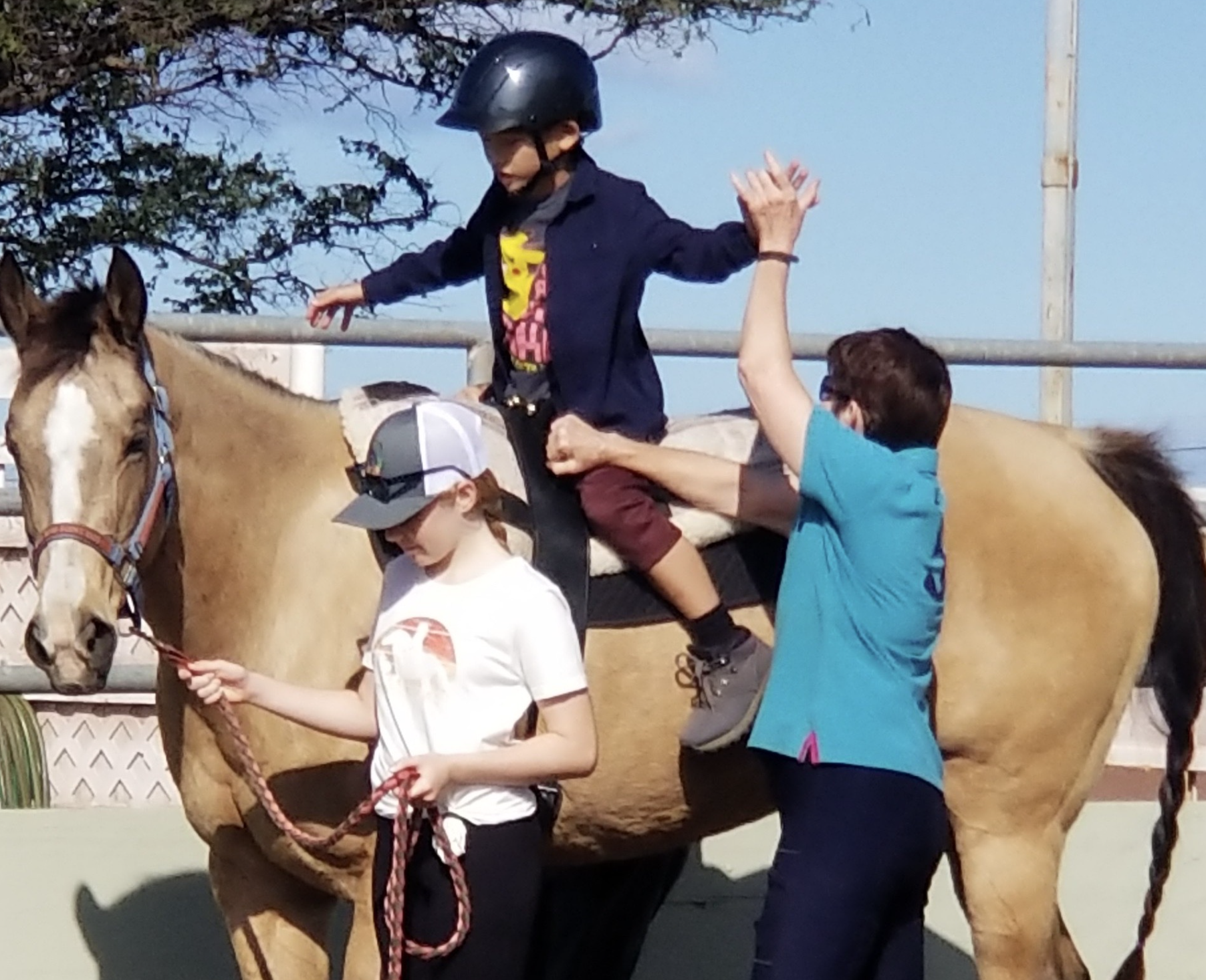 Children learning to ride a horse with guidance from two adults at an outdoor equestrian facility.