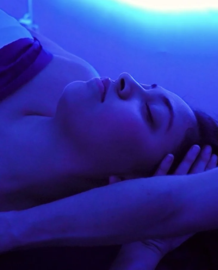 A person lying in bed with closed eyes, resting under blue lighting, with one hand touching the side of their face.