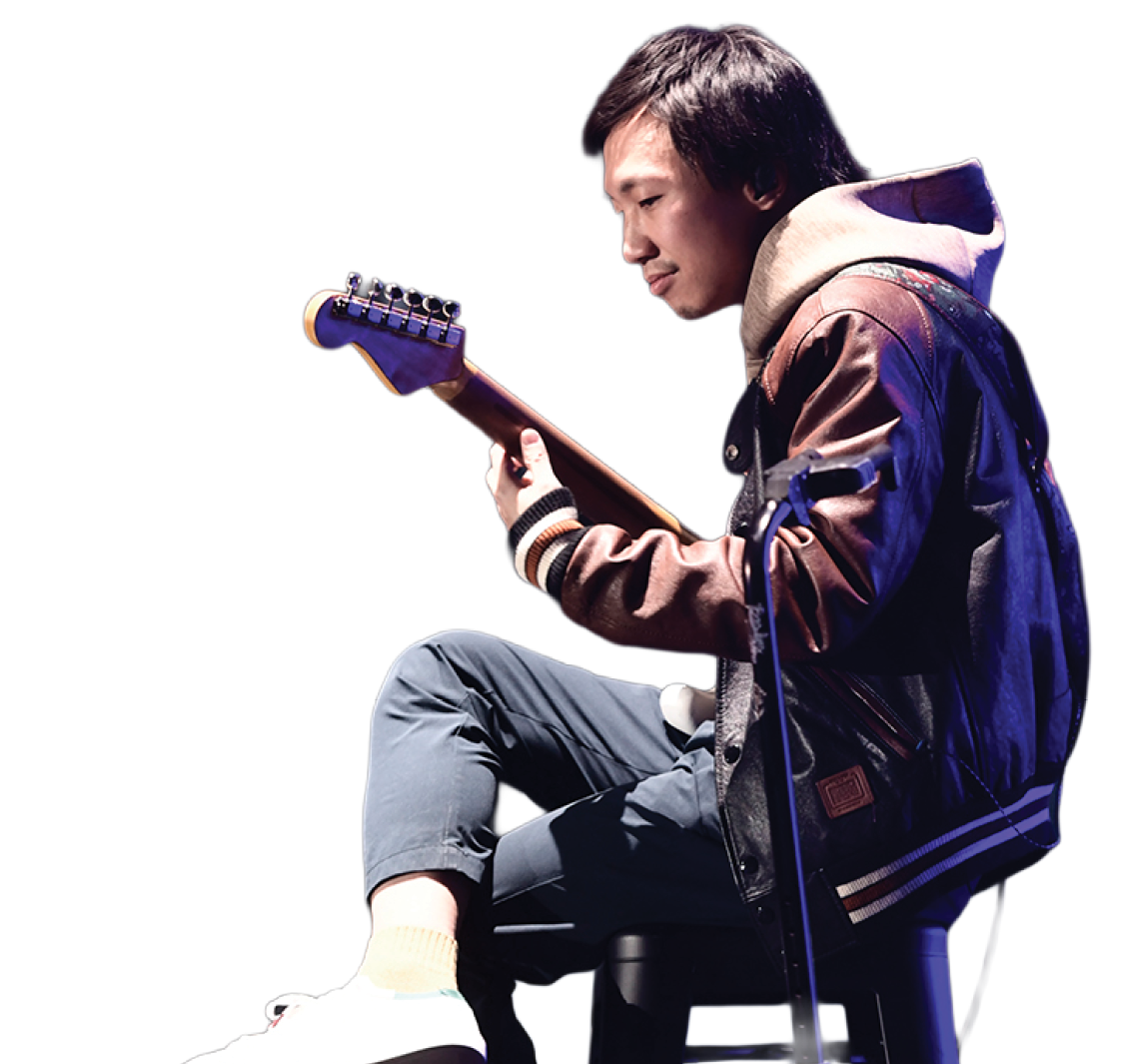 A young man with short dark hair sitting on a black stool, playing an electric guitar. He is wearing a brown leather jacket, beige hoodie, gray pants, and white sneakers with yellow socks. The background is dark. Teriver Cheung