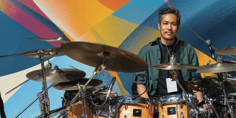 A man sitting behind a drum set with a colorful abstract mural in the background. John Thomas