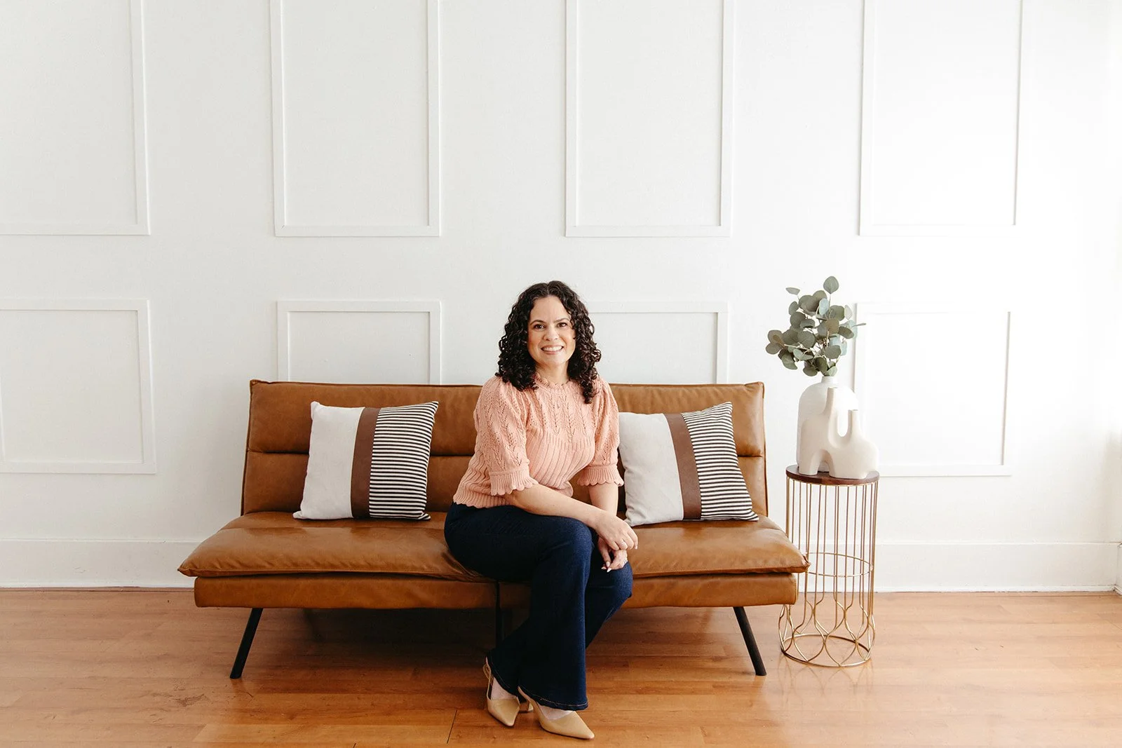 Jaclyn Staubach, LCSW -  anxiety, burnout, and chronic illness therapist sitting on a brown leather sofa in a minimalist living room with white paneled walls, a white decorative vase with greenery on a side table, and light wooden flooring.
