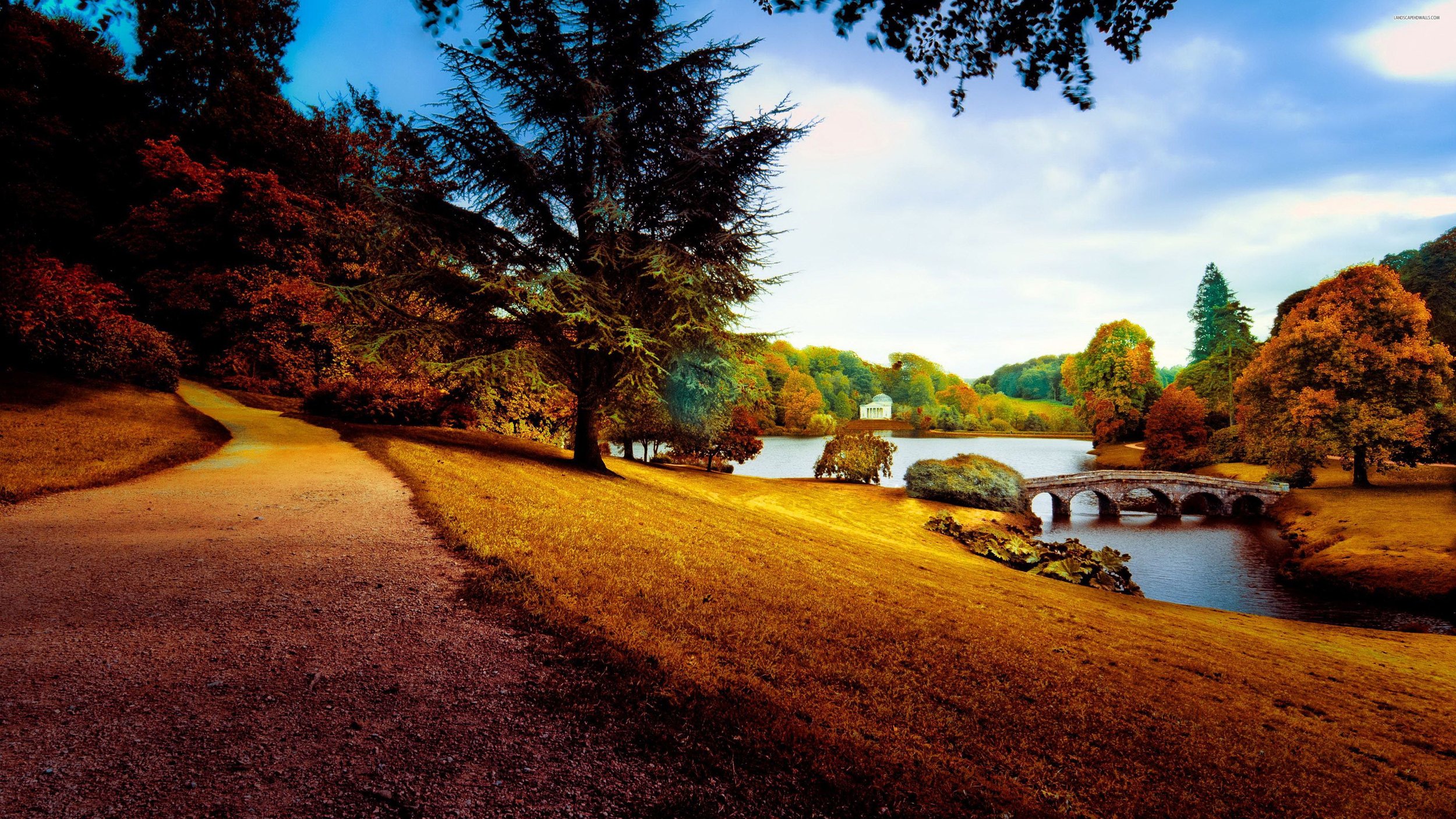 Scenic view of a park with a winding gravel path, colorful autumn trees, a lake, a stone bridge, and a white pavilion in the distance.