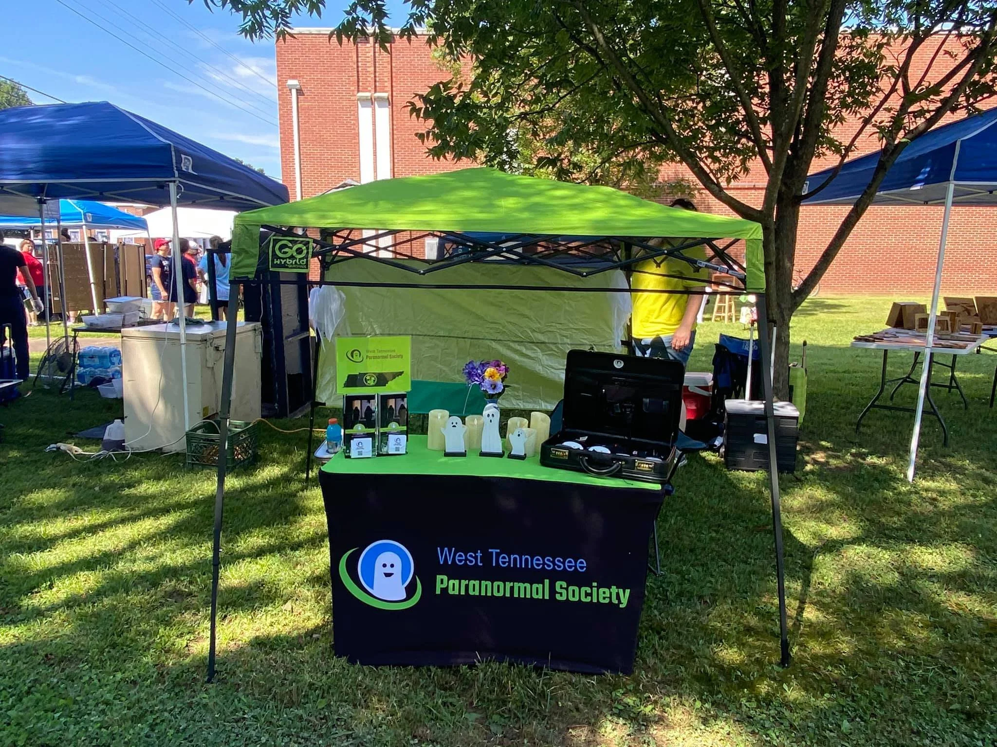 A booth for the West Tennessee Paranormal Society at an outdoor event, featuring a black table with a green and black tablecloth, ghost logo, and various paranormal-themed items on display, set up under a green canopy in a grassy area with other tents and a brick building in the background.