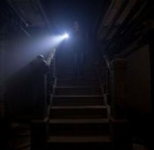 A person holding a flashlight, shining light upward toward a bright light source at the top of a staircase in a dark area.