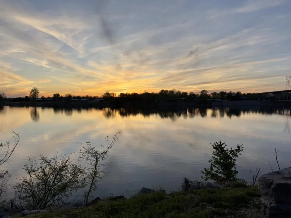 Sunset over a calm lake with trees and a bridge in the distance, reflection of the sky on the water.