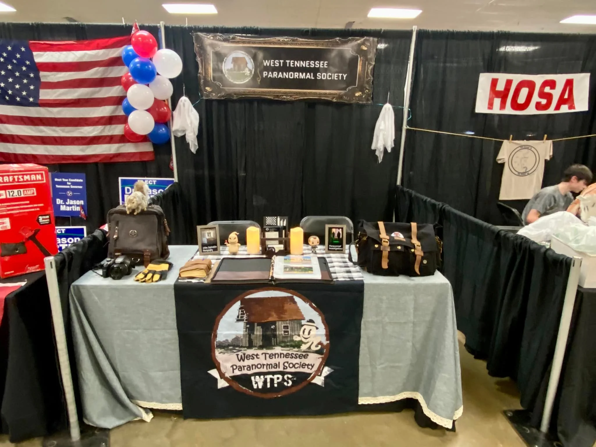 Exhibit booth for West Tennessee Paranormal Society at a convention with American flag, balloons, framed displays, and assorted paranormal equipment.