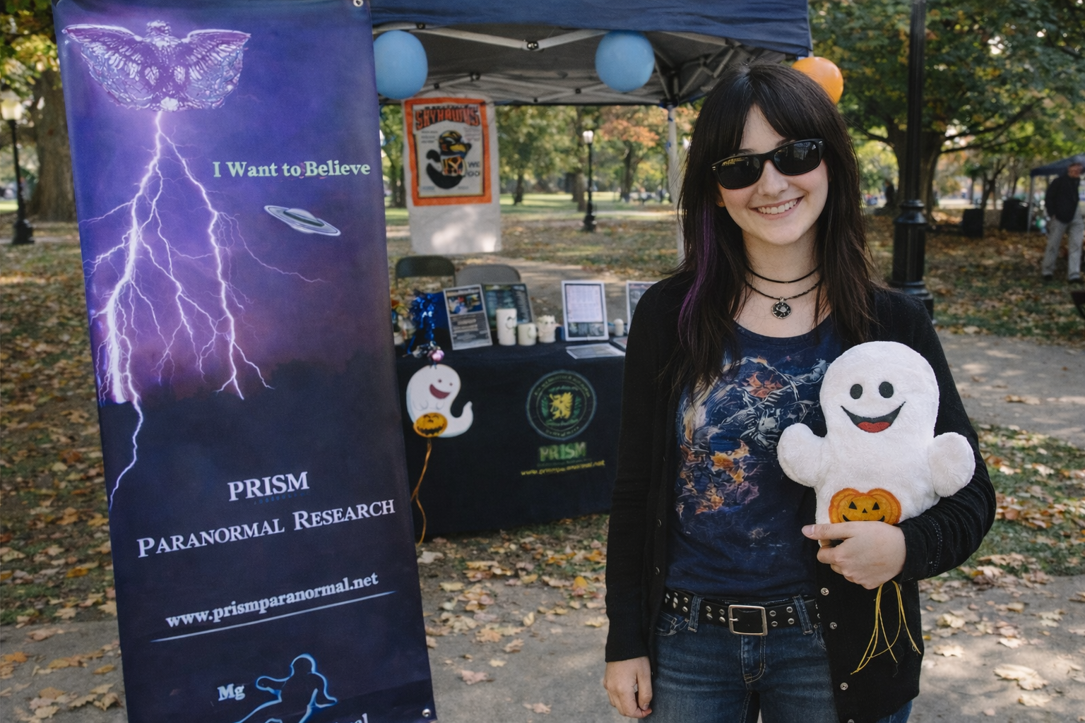 A young woman wearing sunglasses and a semi-goth outfit stands smiling beside a Halloween-themed university booth for the paranormal research and investigation society of martin now known as forgotten places society in savannah,tn.