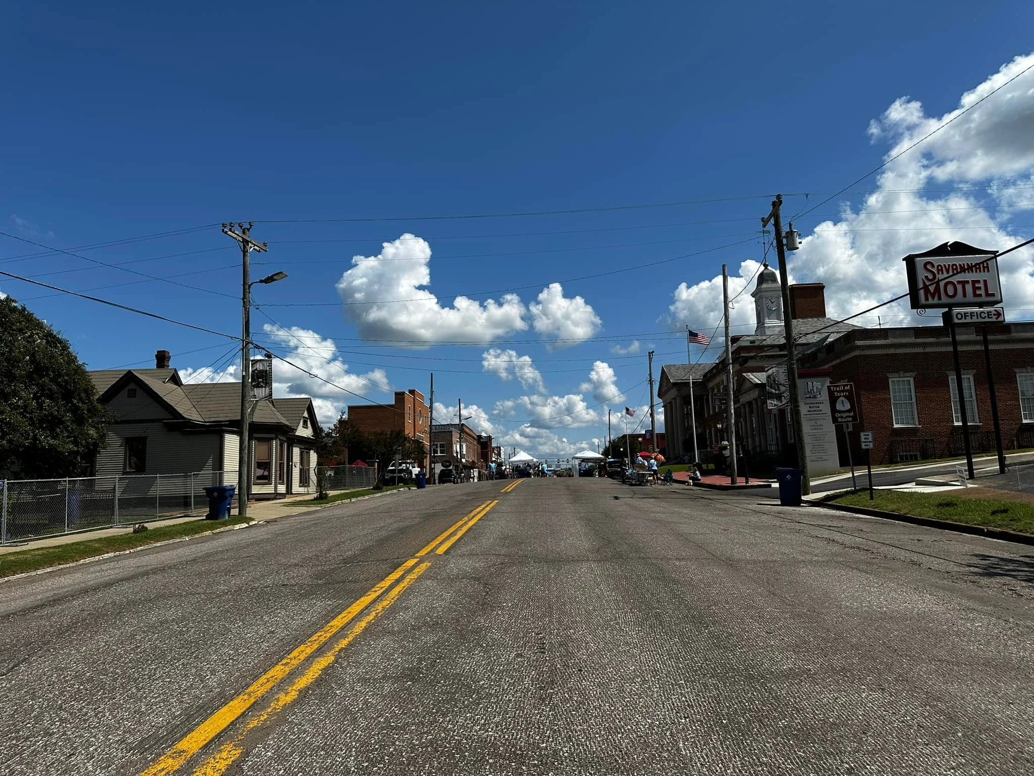 A small town street scene with a clear blue sky and fluffy clouds, featuring a motel and some shops on the right side, and residential houses on the left, with a paved road and yellow dividing lines.