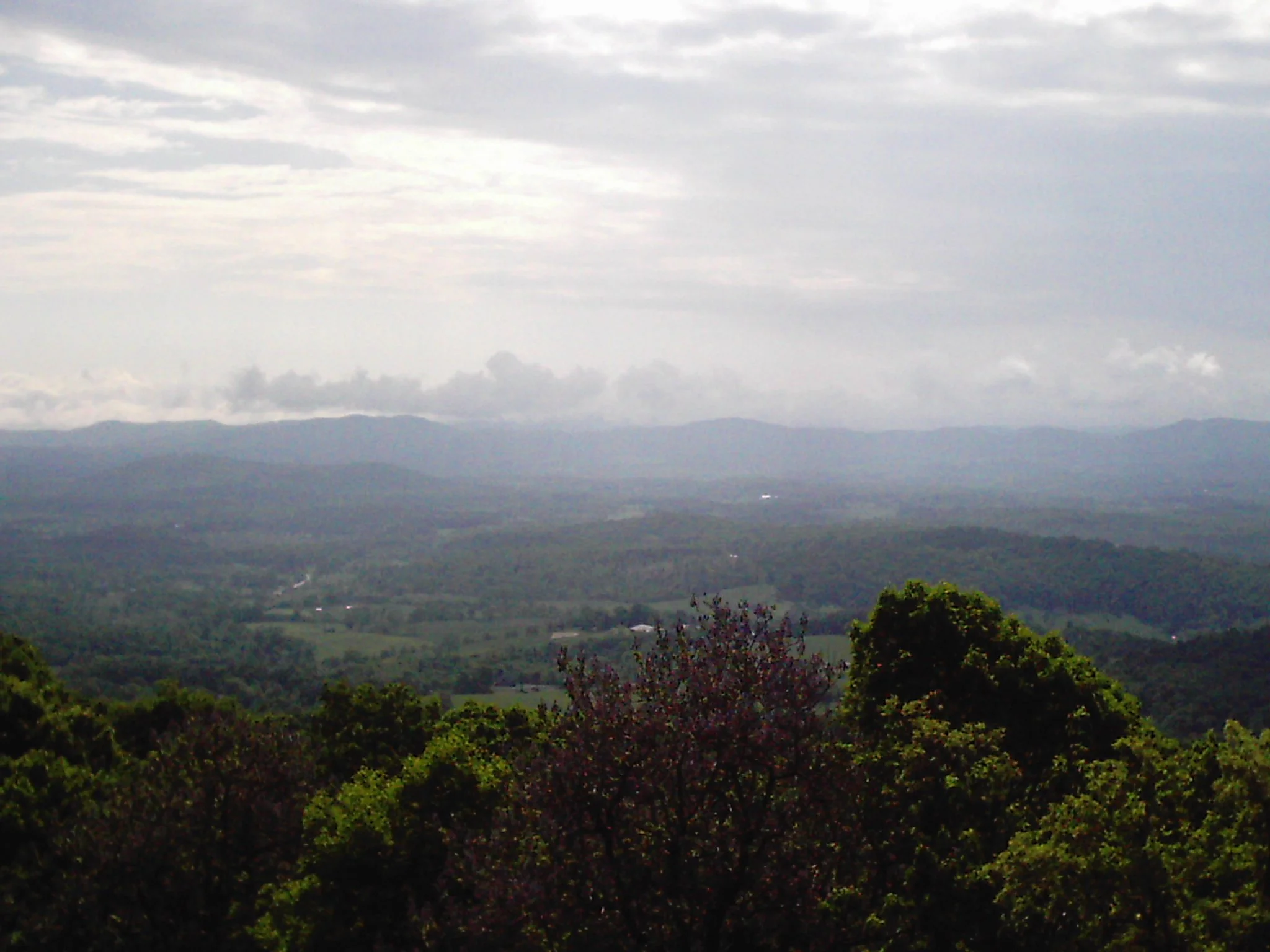 Scenic view of rolling hills and mountains under a cloudy sky, with trees in the foreground.
