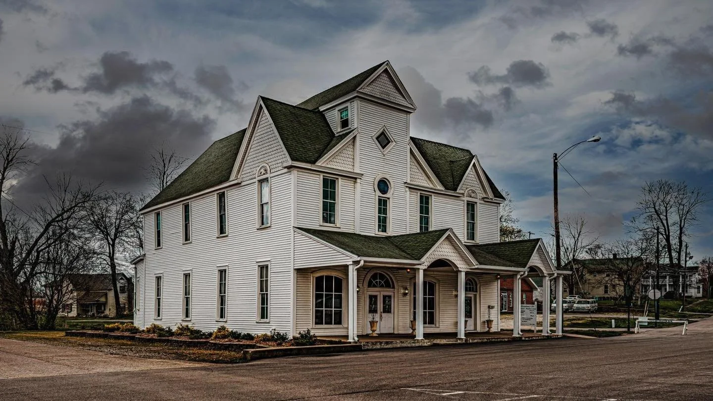A large, white, Victorian-style house with multiple gables, tall windows, and a front porch with columns, set against a cloudy sky.