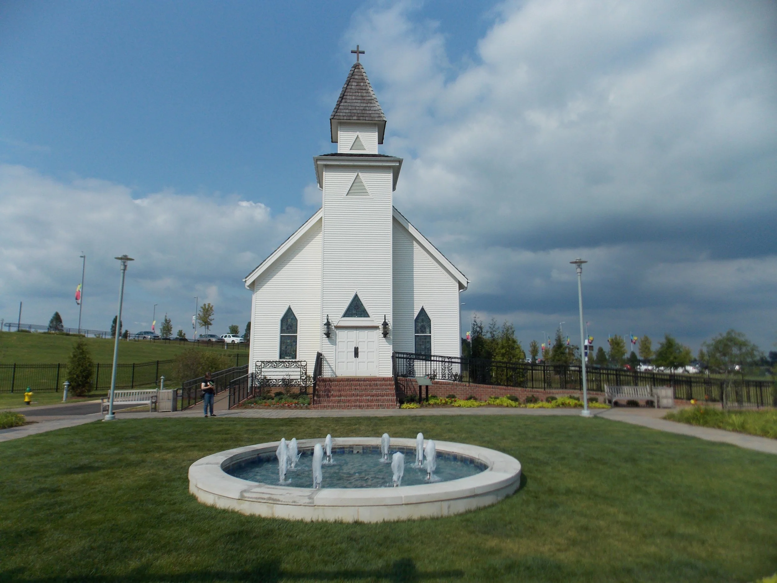 A white church with a steeple, surrounded by a lawn, with a round fountain in the foreground and a partly cloudy sky.