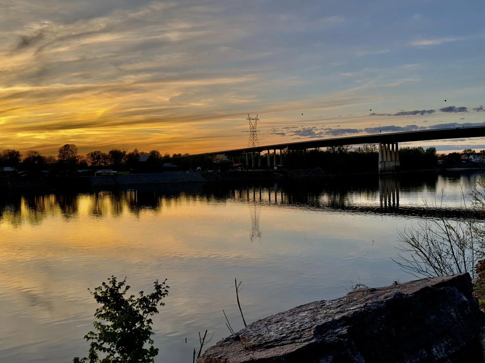 A sunset over a river with a bridge and power lines, calm water reflecting the sky, rocks and trees along the riverbank in the foreground.