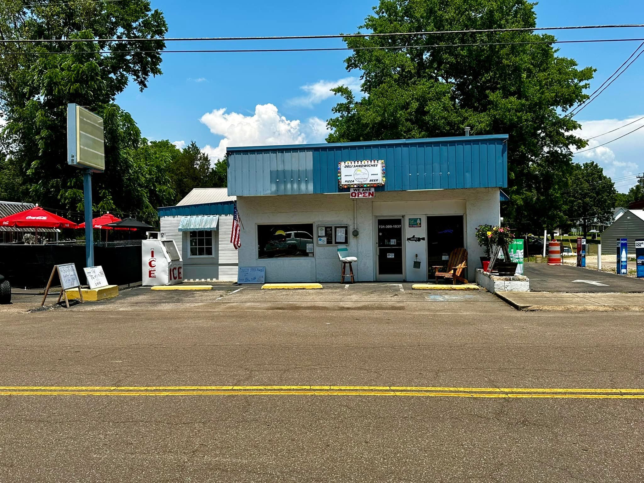 Small roadside sandwich shop with a blue roof, white walls, and a front porch area. There are ice coolers outside, a small wooden Adirondack chair, hanging flower baskets, and outdoor umbrellas with red in the background. Tall trees and a partly clou
