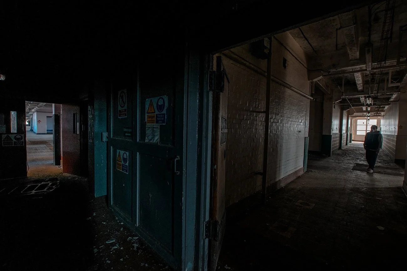 Dark, abandoned hallway with a person walking in the distance, windows letting in some light.
