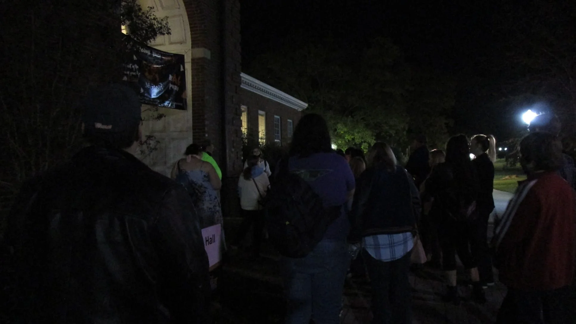 People standing in a line outside at night, with a building and trees in the background, and a light illuminating the scene.