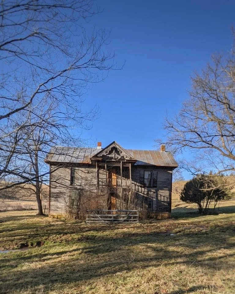 An old two-story wooden house with a metal roof, surrounded by leafless trees and bushes, under a clear blue sky, with a grassy lawn in the foreground.