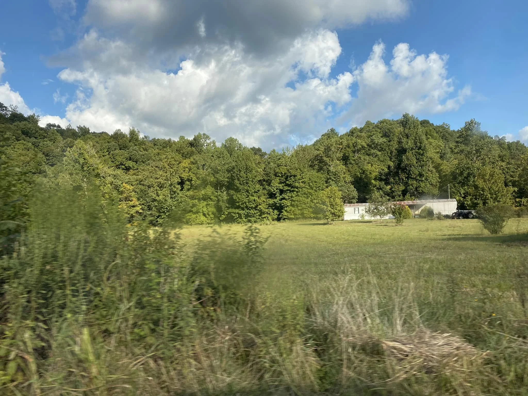 A rural landscape with a grassy field, trees in the background, a small building, and a few parked vehicles under a partly cloudy sky.