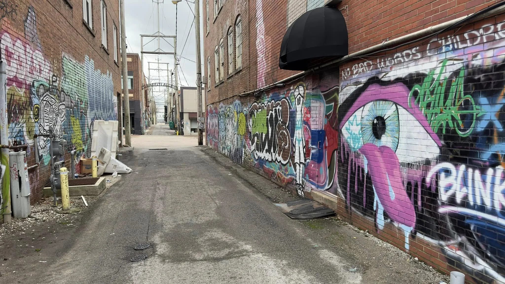 An alleyway with brick buildings on either side covered in colorful graffiti art, including a large eye with pink eyelids and dripping tears on the right wall.