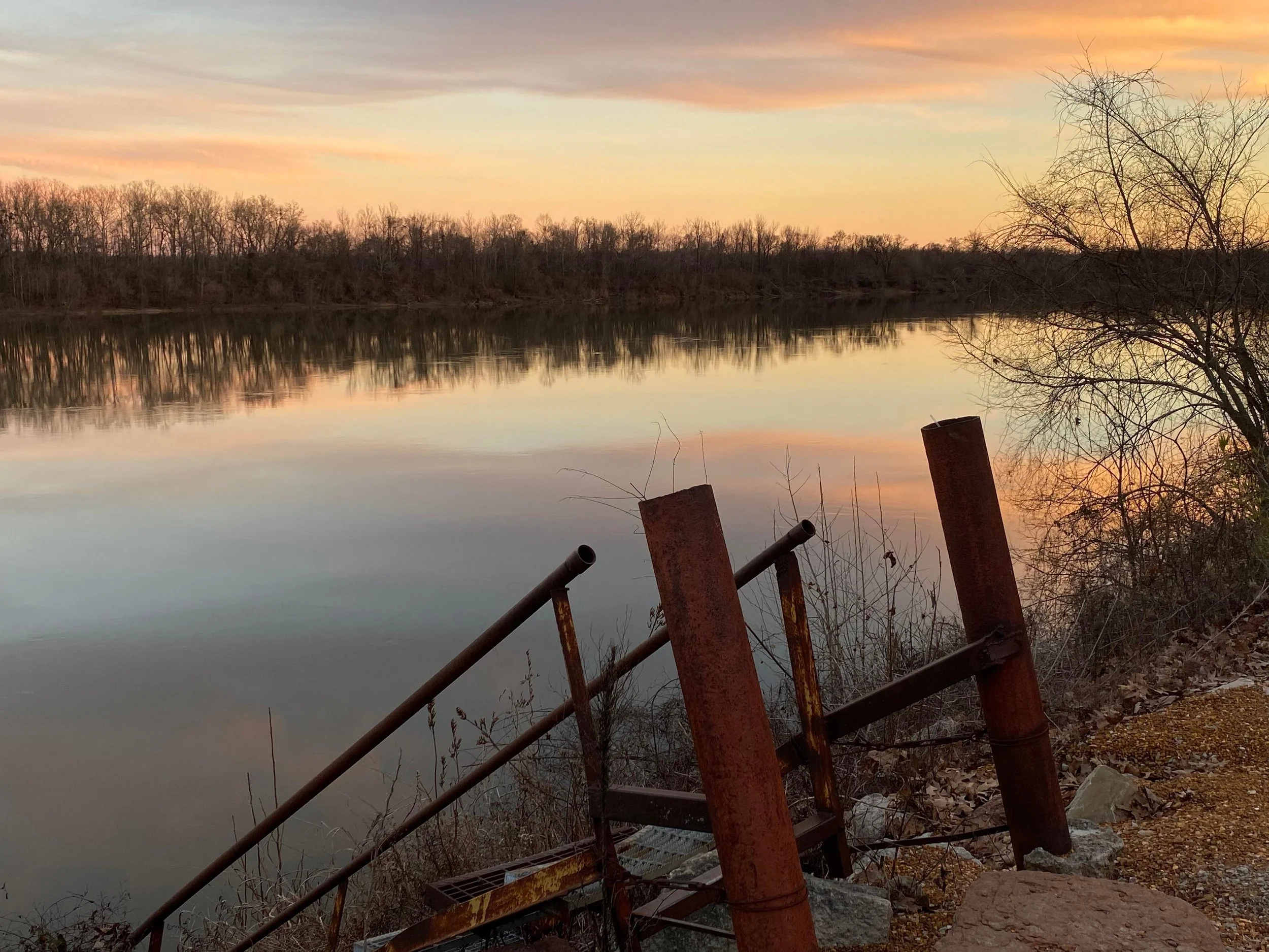 A calm lake reflecting a colorful sunset sky with trees along the shoreline and rusted metal railings in the foreground.