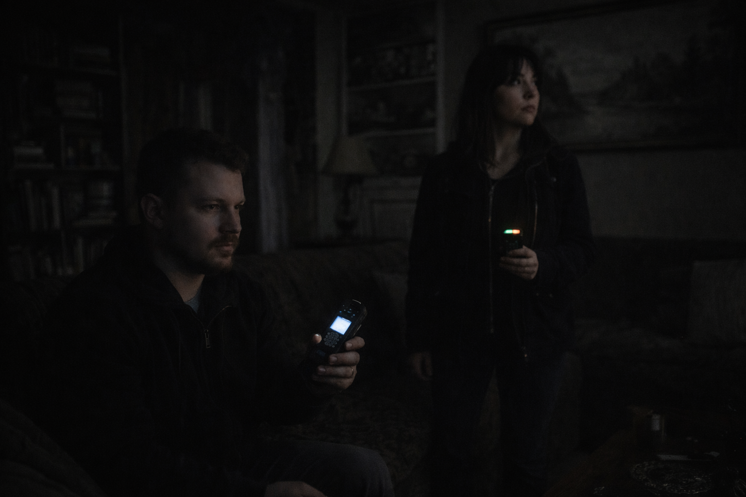 A man and woman are sitting and standing in a dark room, both holding cell phones with screens lit. The man is sitting on a couch, looking at his phone, while the woman stands nearby, looking elsewhere, also holding her phone.