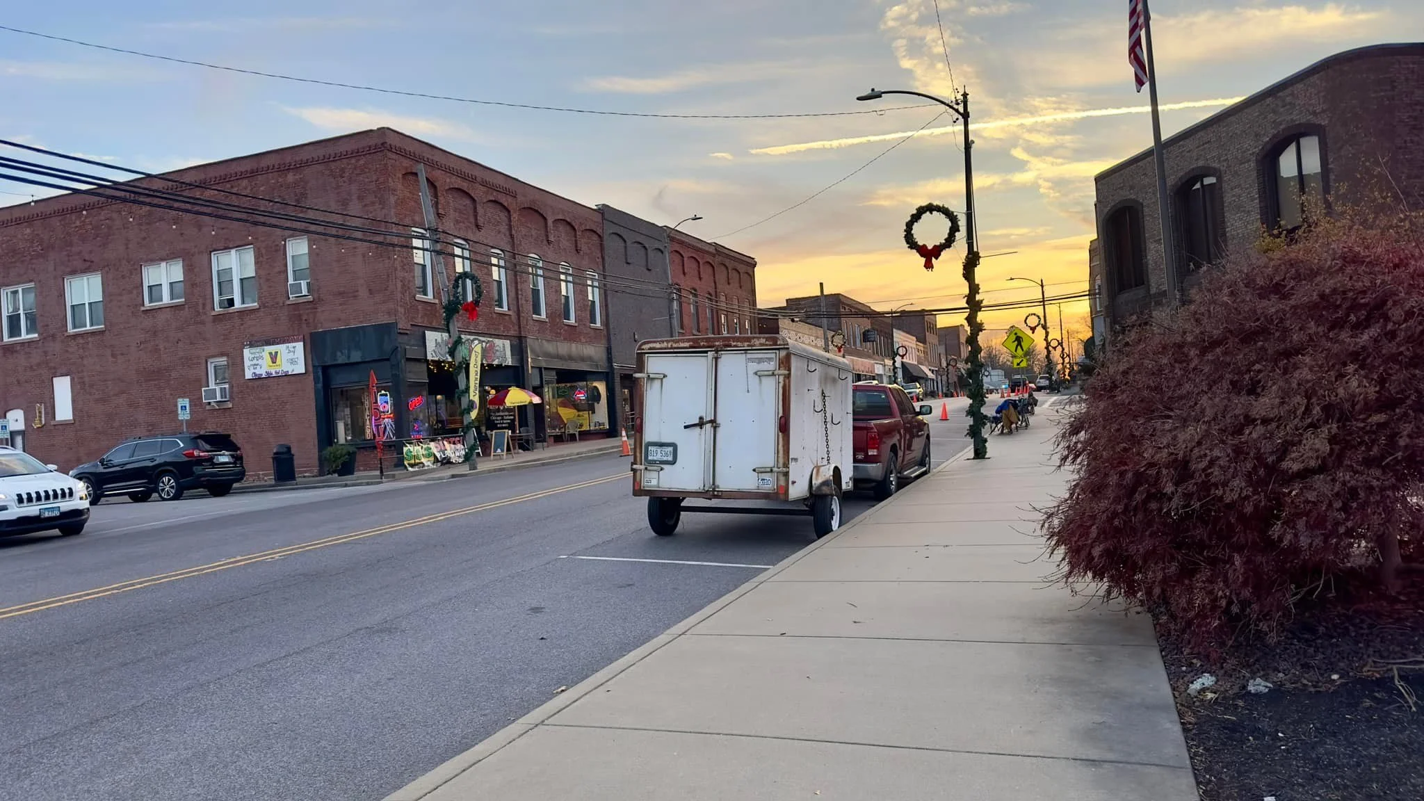 A small trailer attached to a red pickup truck parked on the side of a street in a small town, with shops and buildings decorated for the holidays