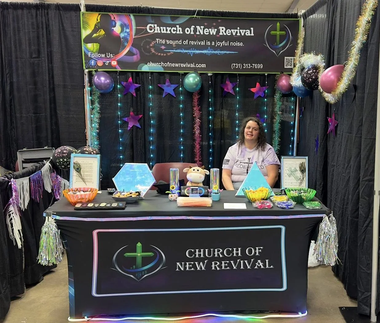 A promotional booth for the Church of New Revival decorated with balloons, star-shaped and tinsel decorations, soft lighting, and a banner at the top. A woman sits behind the table, which displays candy bowls, framed artwork, and a plush cow toy, with a black tablecloth featuring the church's logo and name.
