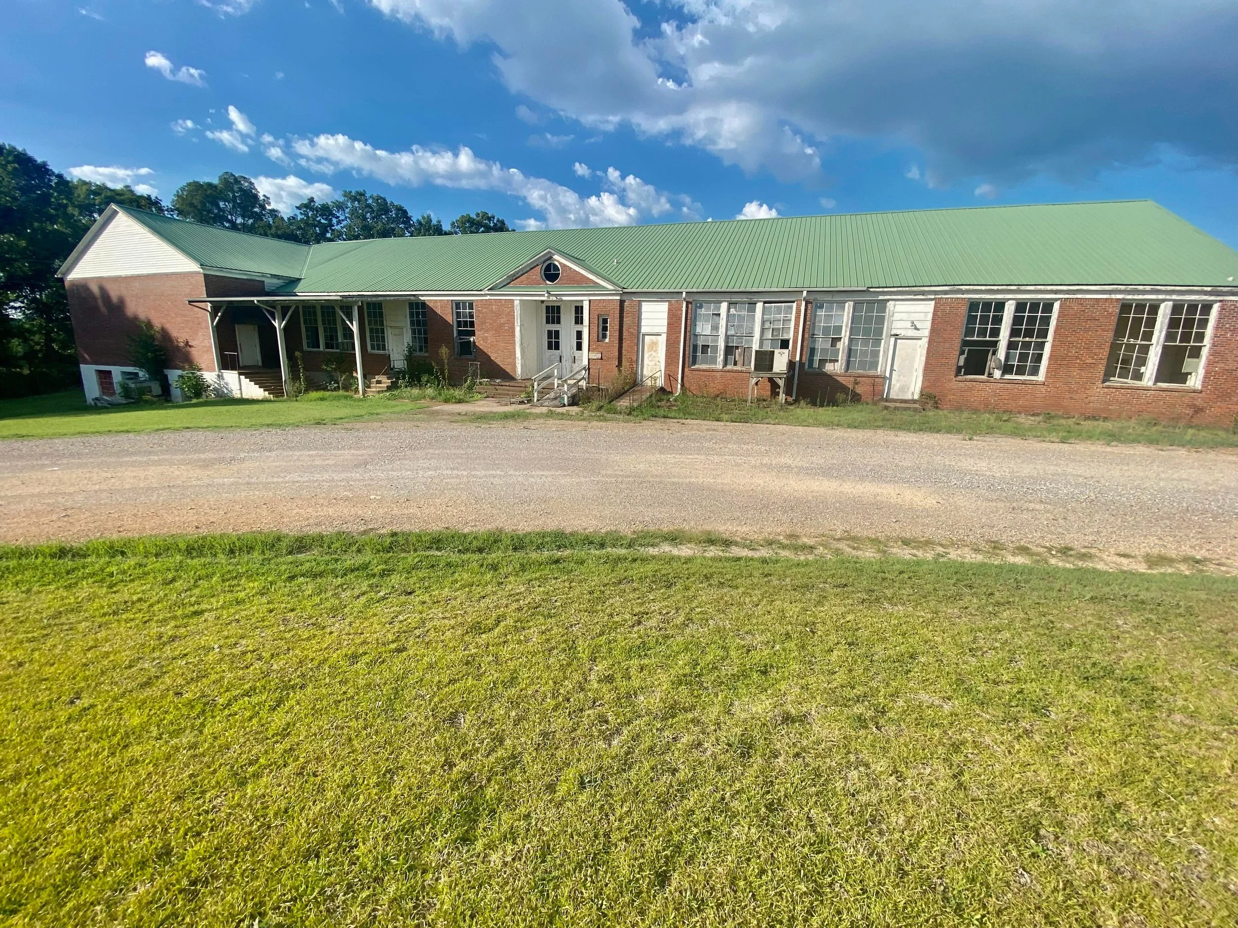 A long, one-story brick building with a green metal roof and multiple windows. Some windows are open, with window units installed. There are doors and small porches along the front. The building is surrounded by a grassy area and a gravel driveway, with a backdrop of trees and a partly cloudy sky.