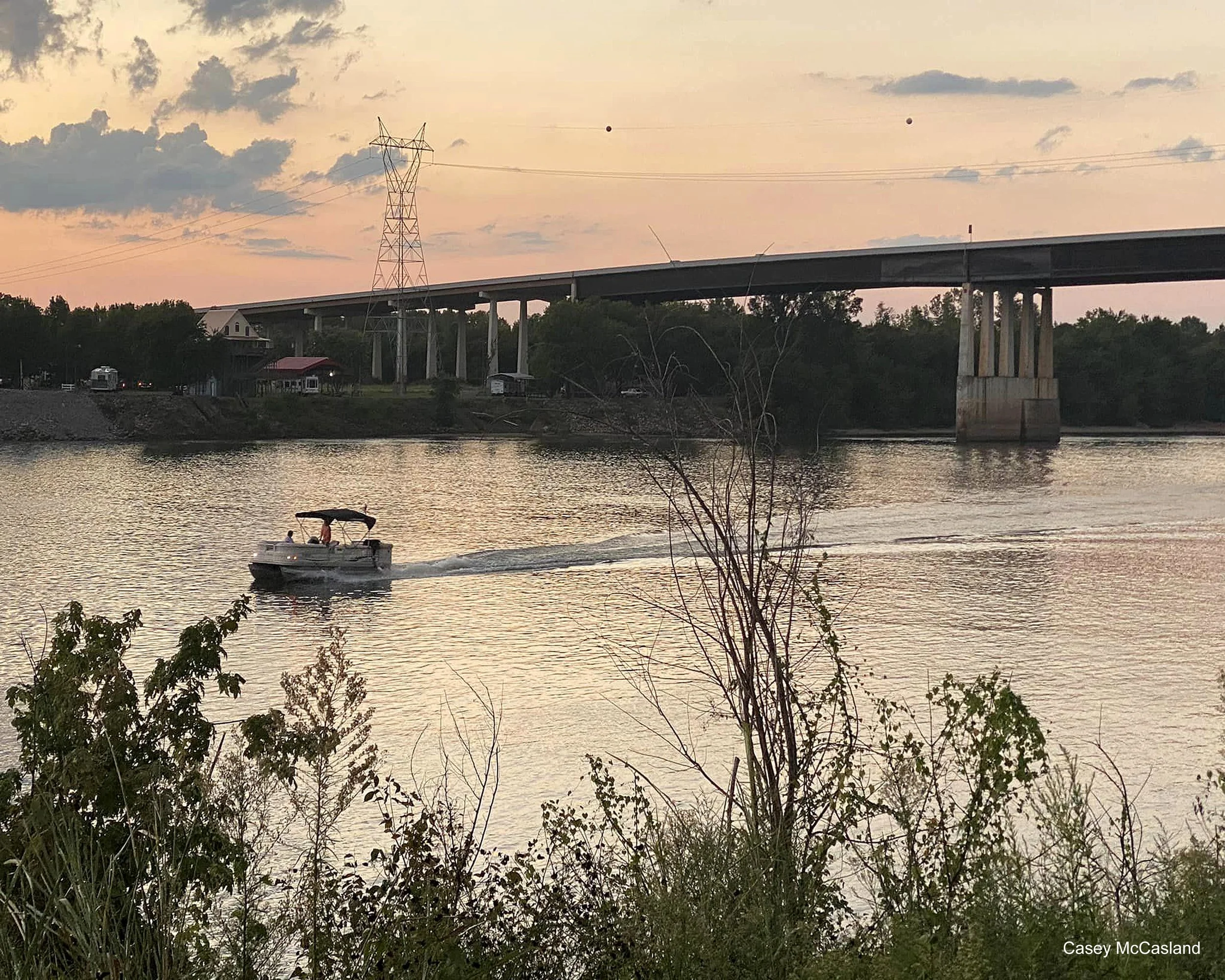 A boat with two people on a river during sunset, with a large bridge, trees, and a partly cloudy sky in the background.