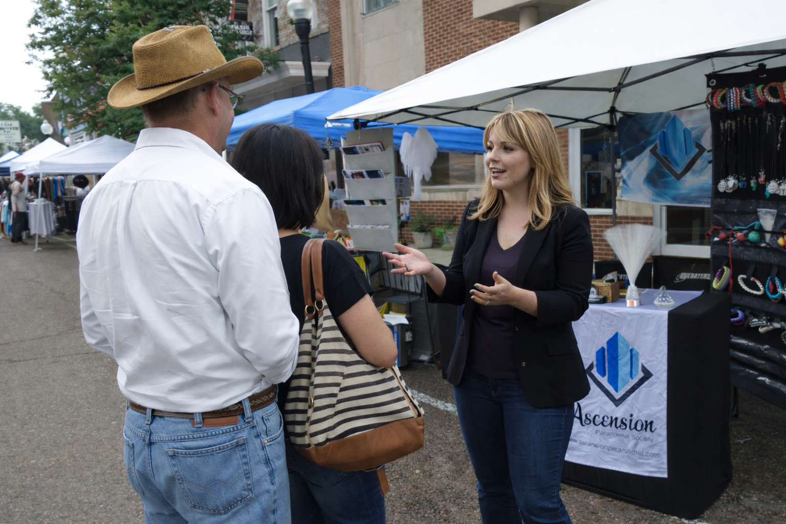 A woman is talking to two people at an outdoor market booth, with stalls and tents visible in the background.