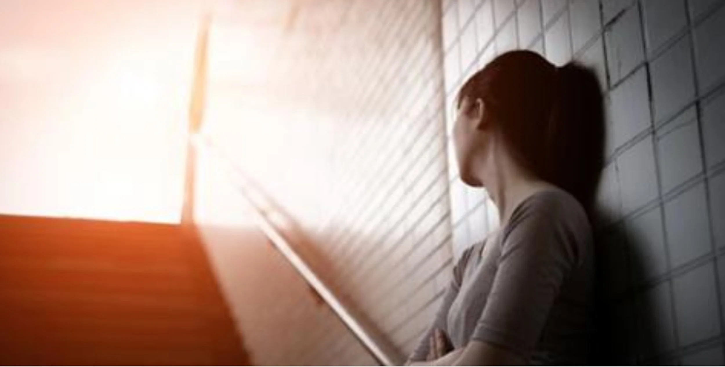 A woman with long dark hair sitting against a tiled wall, looking away, with sunlight coming through window blinds.