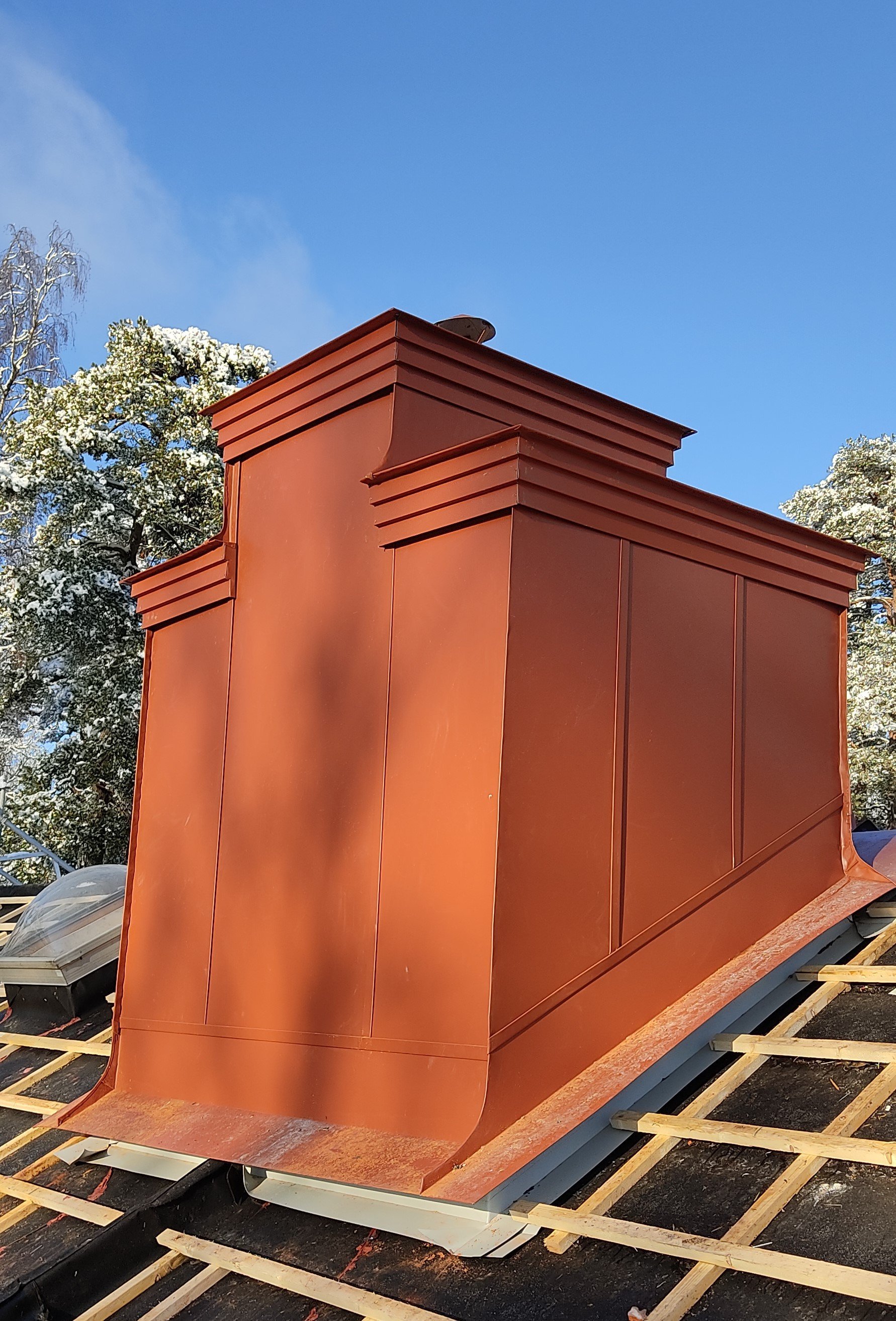 En röd plåttakshatt på ett tak under renovering, omgiven av byggnadsramar och träd med snö på.