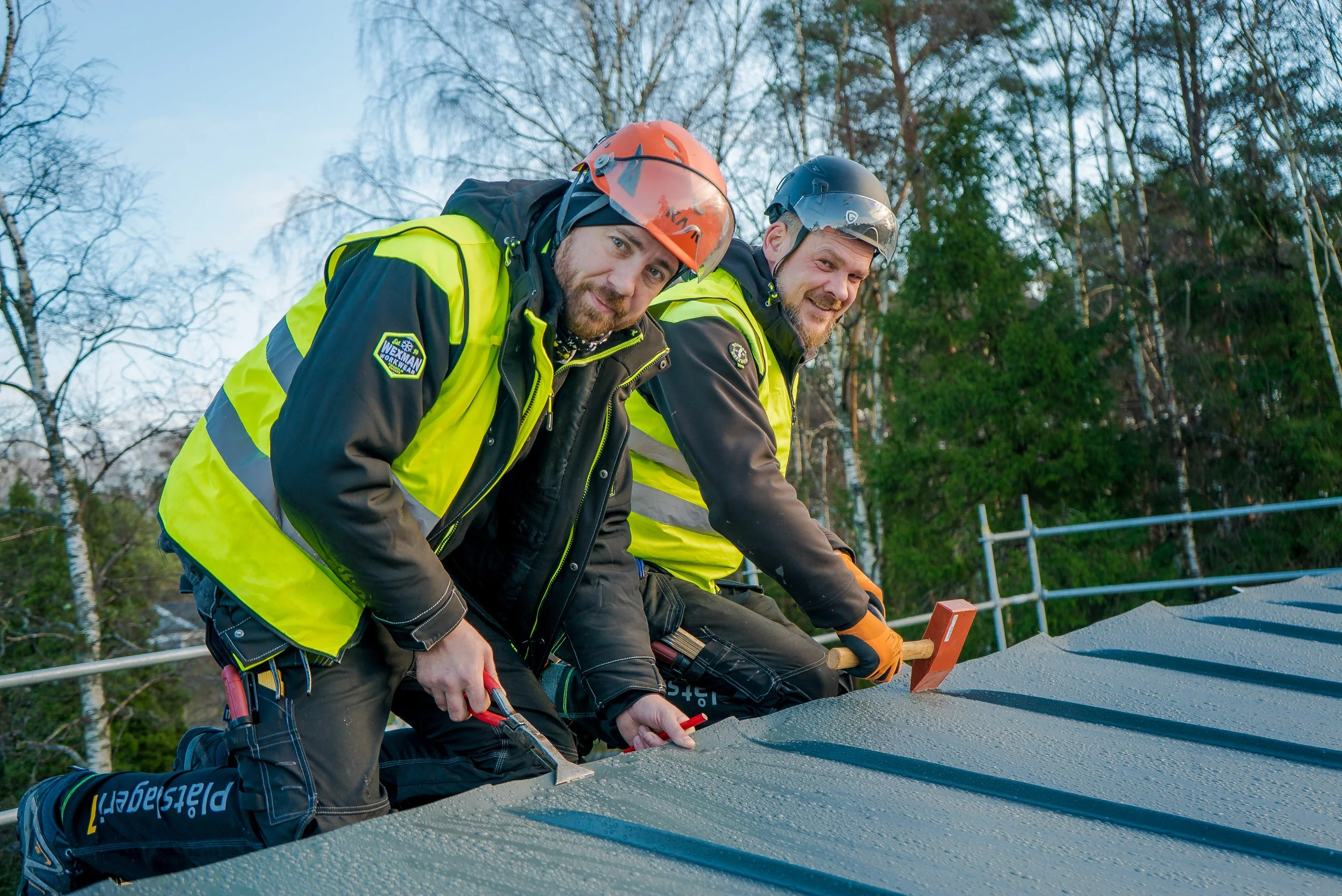 Två män i säkerhetsutrustning arbetar på ett tak, med skog i bakgrunden.
