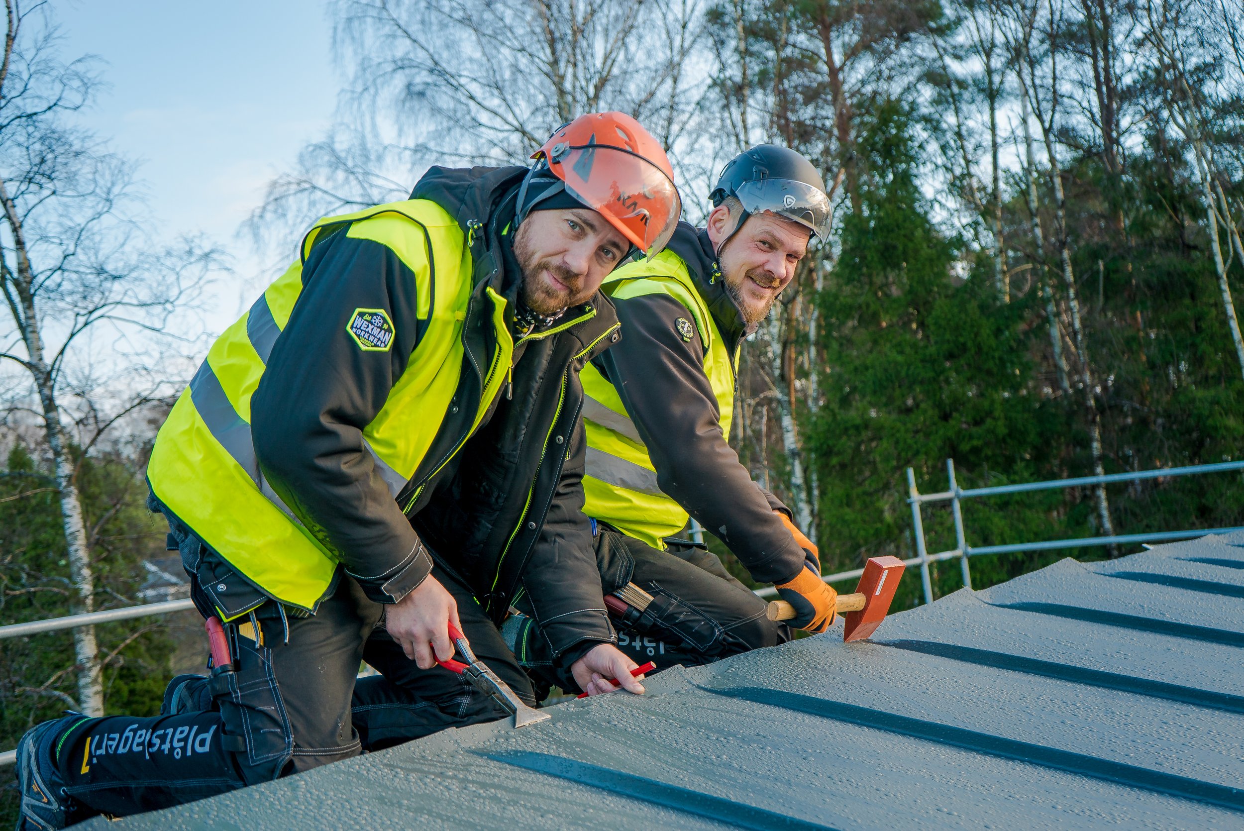 Två män i säkerhetsutrustning arbetar på ett tak, med skog i bakgrunden.