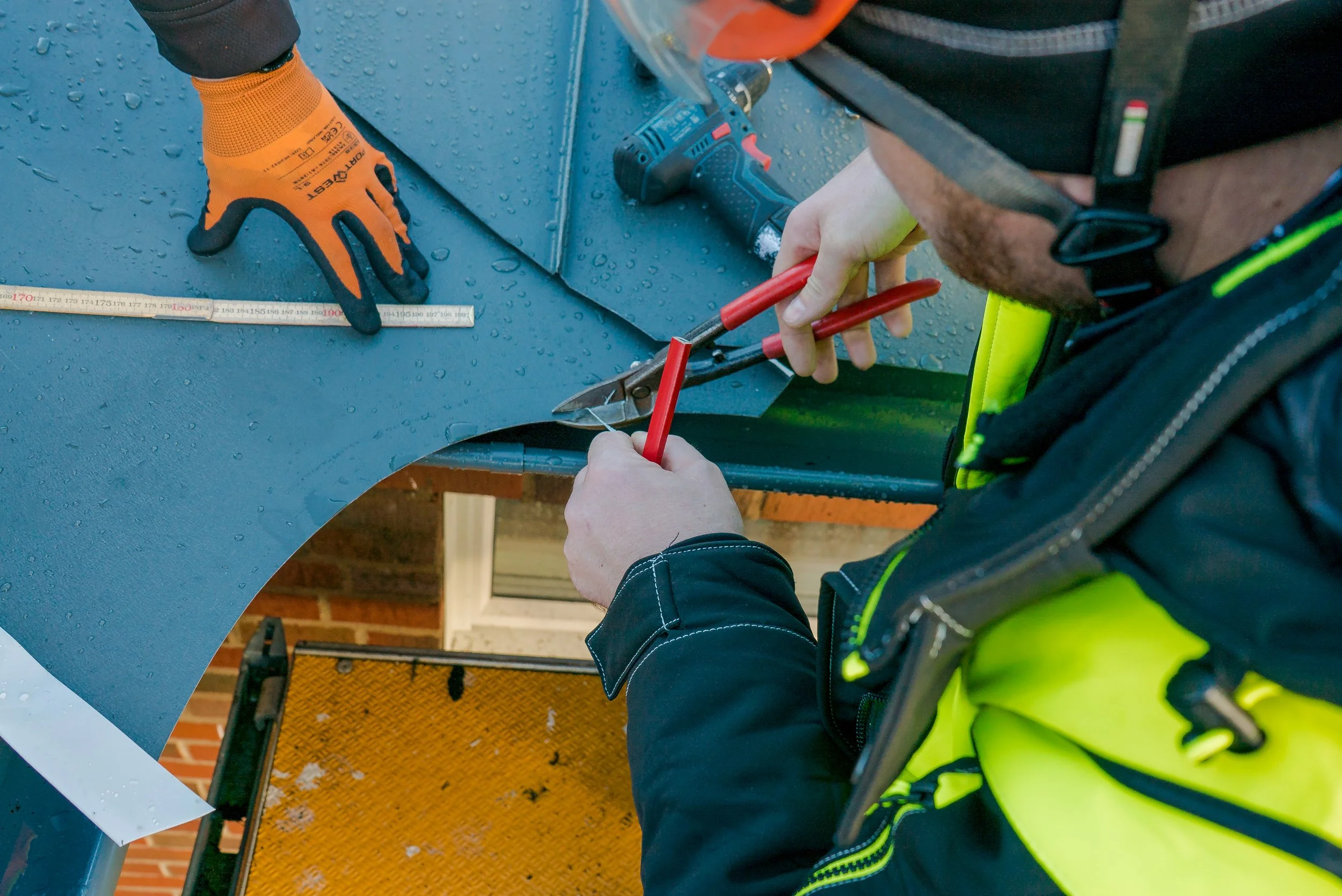 Person som arbetar med metall, använder verktyg för att böja eller forma metallen, med ett annat par händer som håller metallbiten på plats. Händerna har skyddshandskar, och personen bär en reflexväst.