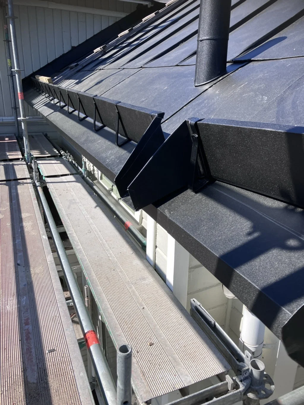 Modernt svart tak med ventilationsventil och rostfria skorstensskydd, byggnadsställningar i förgrunden.