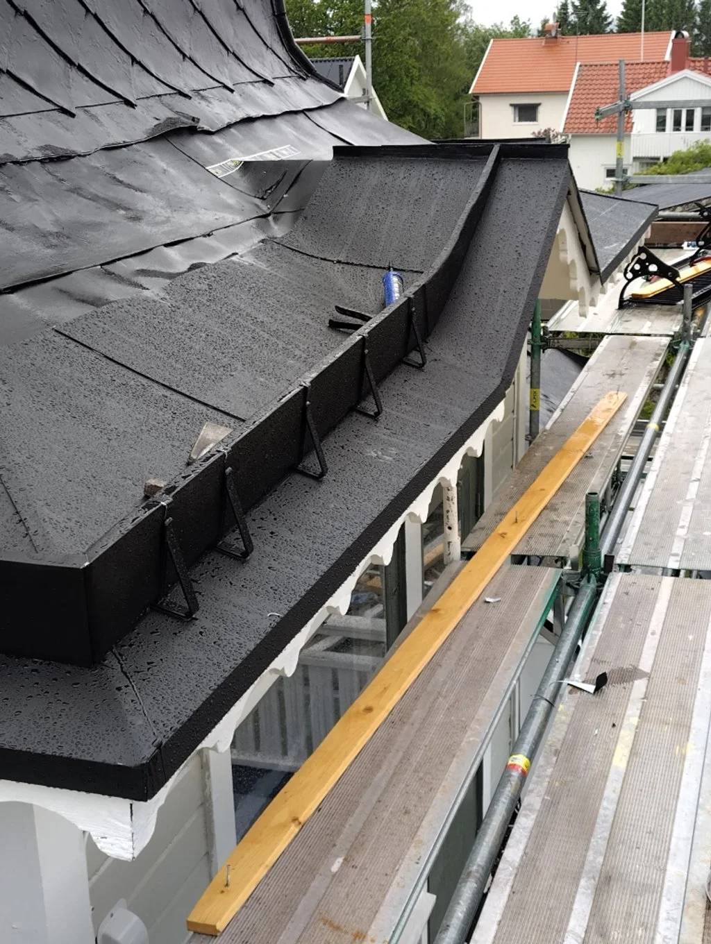 Takrenovering pågår på ett hus med takplåt och dynor, utsikt över grannhus och träd.